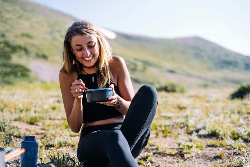 woman in black tank top and black pants sitting on ground holding blue ceramic mug during a mindful eating session on a hike